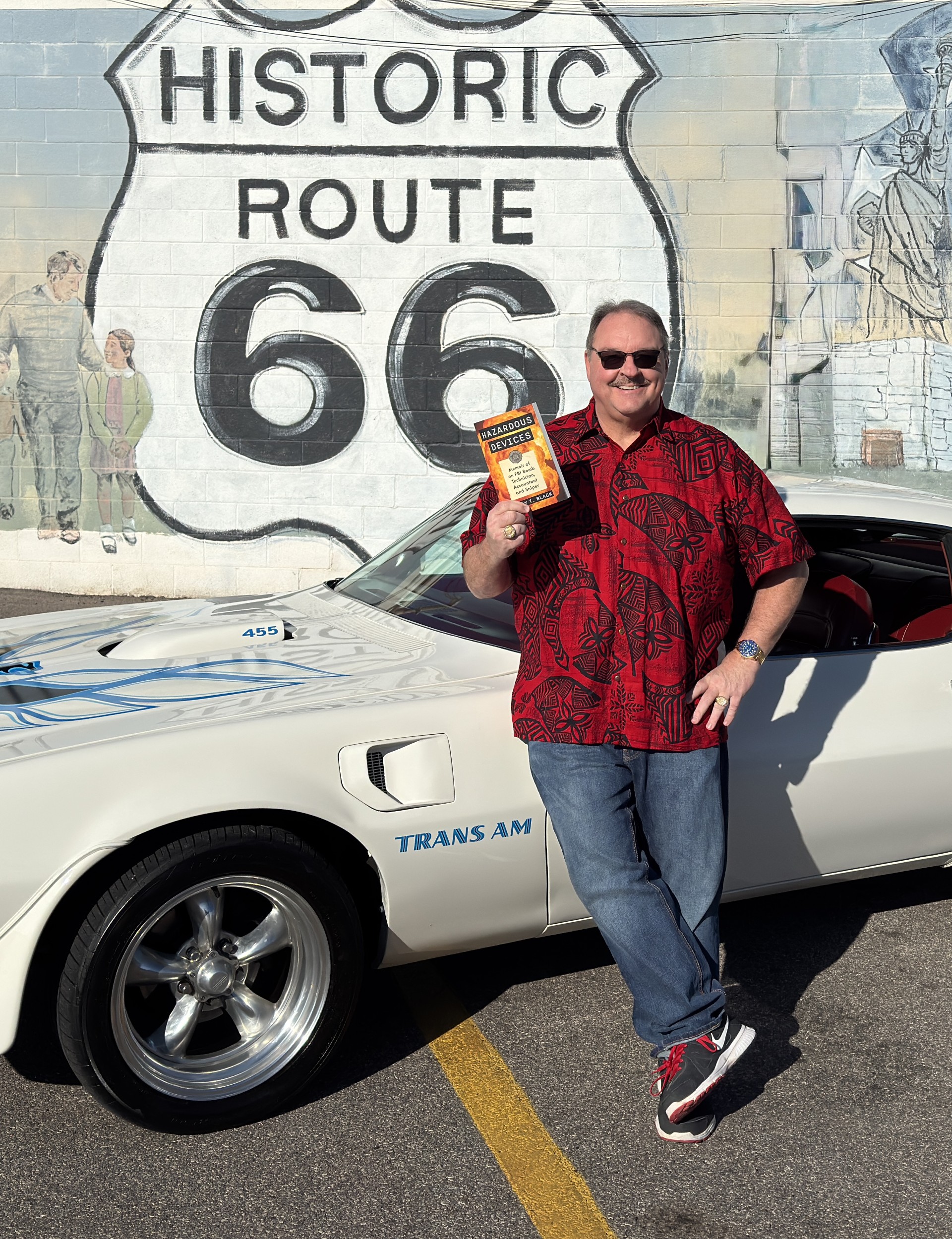 Barry Black is wearing the South Pacific Tribal High Seas Hawaiian shirt, posing with his book "Hazardous Devices" in front of his '73 Firebird Trans Am